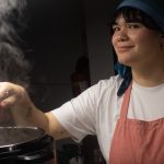 Chef paraense sorrindo enquanto cozinha, com vapor subindo de uma panela.
