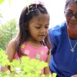 Criança e idosa em projeto social, com plantas ao fundo.
