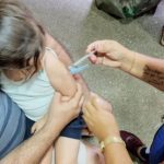 Criança recebe vacina no braço, com ajuda do pai e profissional de saúde.