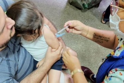 Criança recebe vacina no braço, com ajuda do pai e profissional de saúde.