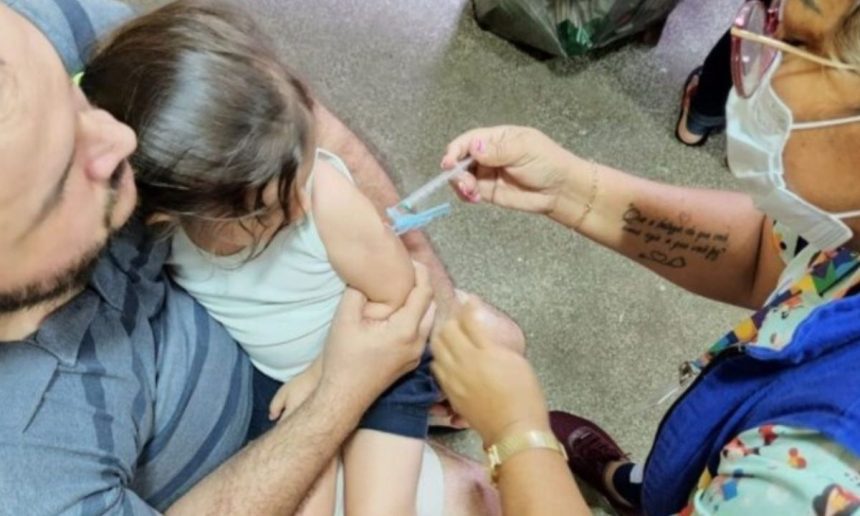 Criança recebe vacina no braço, com ajuda do pai e profissional de saúde.
