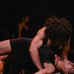 Dançarinos performam em palco escuro, com foco na interação e expressão corporal.