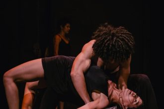 Dançarinos performam em palco escuro, com foco na interação e expressão corporal.