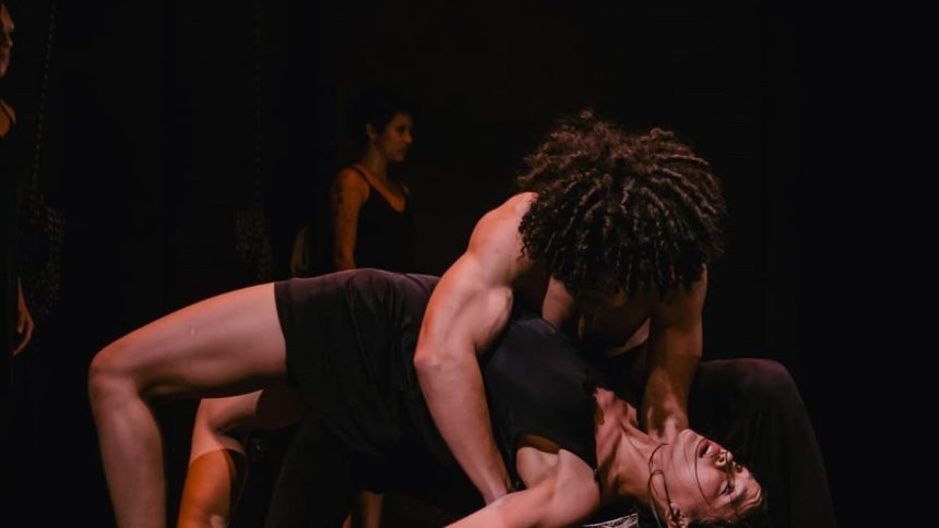 Dançarinos performam em palco escuro, com foco na interação e expressão corporal.