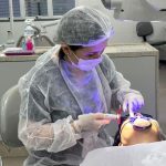 Dentista realizando procedimento odontológico em paciente, usando equipamento e vestindo equipamentos de proteção.