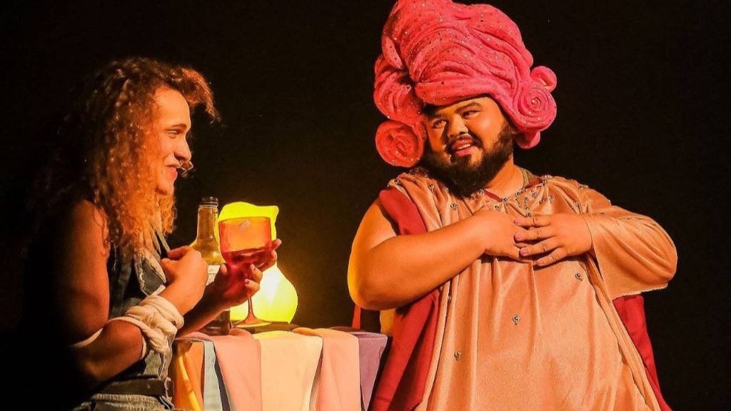 Dois atores em figurinos elaborados, em cena de peça teatral, com luz baixa e atmosfera dramática.