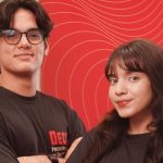 Dois jovens sorrindo em frente a um fundo vermelho com linhas curvas.