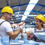 Dois trabalhadores comemoram em fábrica usando capacetes e óculos de proteção.