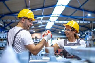 Dois trabalhadores comemoram em fábrica usando capacetes e óculos de proteção.