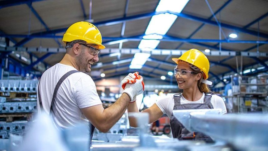 Dois trabalhadores comemoram em fábrica usando capacetes e óculos de proteção.