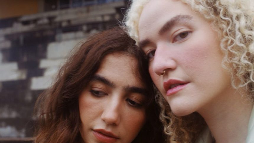 Duas mulheres juntas, uma com cabelo castanho e outra loira, com um fundo desfocado.