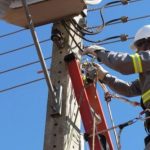 Eletricista trabalhando em poste de energia, com céu azul ao fundo.