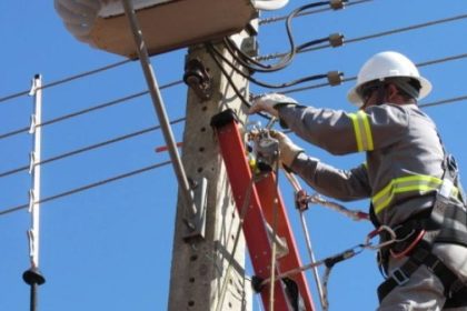 Eletricista trabalhando em poste de energia, com céu azul ao fundo.