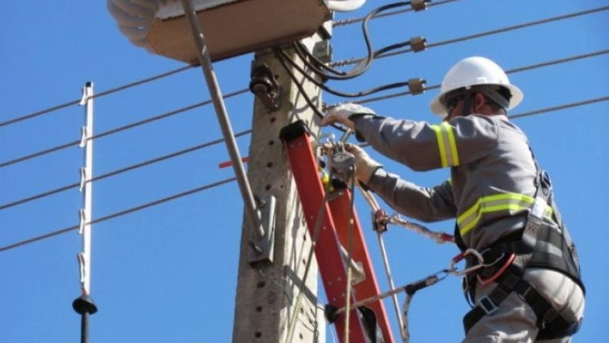Eletricista trabalhando em poste de energia, com céu azul ao fundo.