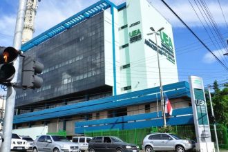 Fachada da Universidade do Estado do Amazonas (UEA) em dia ensolarado, com carros na rua e sinal de trânsito.