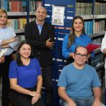 Grupo de pessoas em biblioteca, possivelmente professores ou pesquisadores, com livros e prateleiras ao fundo.