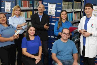 Grupo de pessoas em biblioteca, possivelmente professores ou pesquisadores, com livros e prateleiras ao fundo.