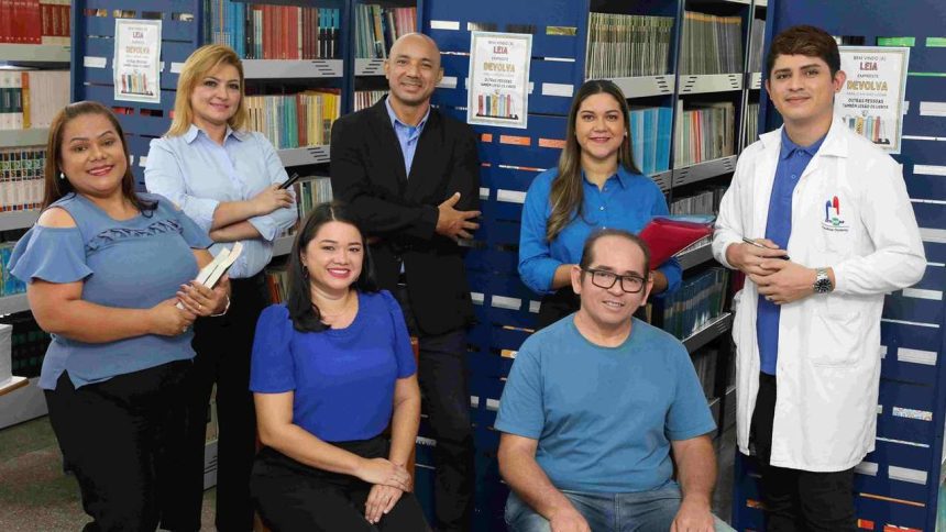 Grupo de pessoas em biblioteca, possivelmente professores ou pesquisadores, com livros e prateleiras ao fundo.