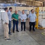 Grupo de pessoas em fábrica, possivelmente uma planta da Collpack, com produtos embalados ao fundo.