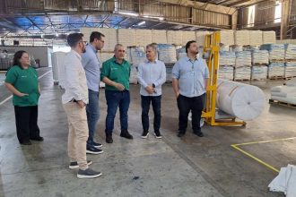 Grupo de pessoas em fábrica, possivelmente uma planta da Collpack, com produtos embalados ao fundo.