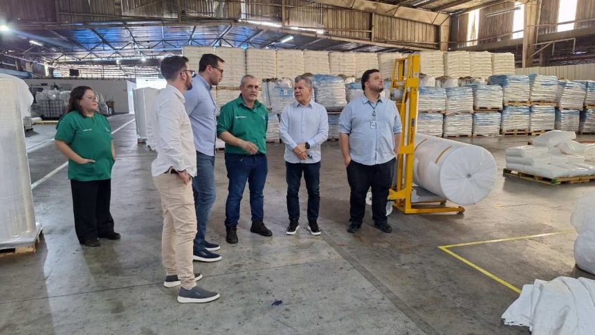 Grupo de pessoas em fábrica, possivelmente uma planta da Collpack, com produtos embalados ao fundo.