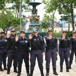 Grupo de policiais femininas em uniforme posando em frente a um chafariz, com mãos levantadas.