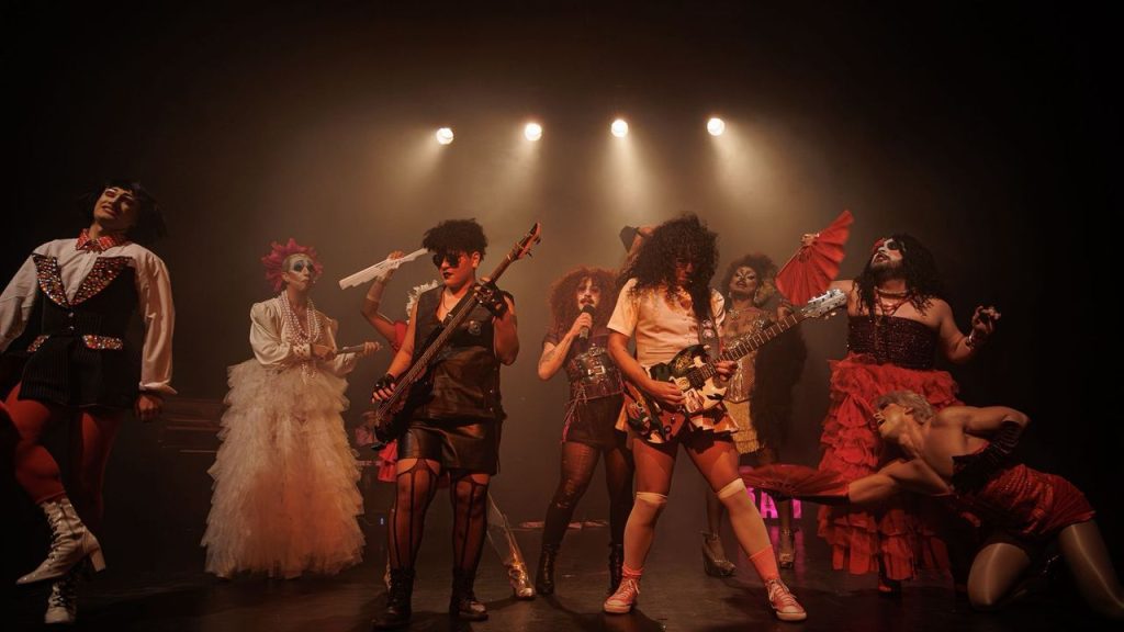 Grupo de teatro performando em palco com luzes, drag queens e instrumentos musicais.