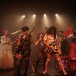 Grupo de teatro performando em palco escuro, com figurinos extravagantes e luzes