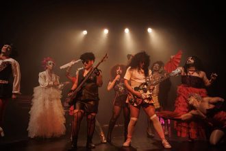 Grupo de teatro performando em palco escuro, com figurinos extravagantes e luzes
