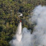 Helicóptero combatendo incêndio florestal na Amazônia, com fumaça e mata densa ao fundo.