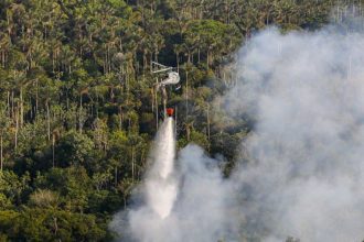 Helicóptero combatendo incêndio florestal na Amazônia, com fumaça e mata densa ao fundo.