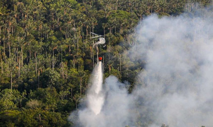 Helicóptero combatendo incêndio florestal na Amazônia, com fumaça e mata densa ao fundo.