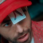 Homem com boné vermelho e carta de baralho no rosto, em evento de cinema.