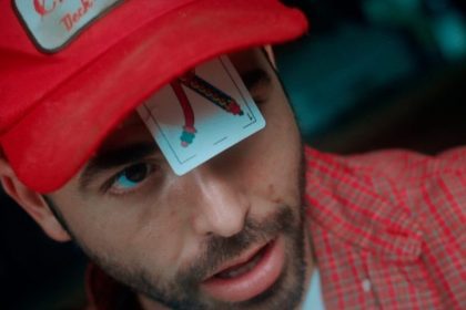 Homem com boné vermelho e carta de baralho no rosto, em evento de cinema.