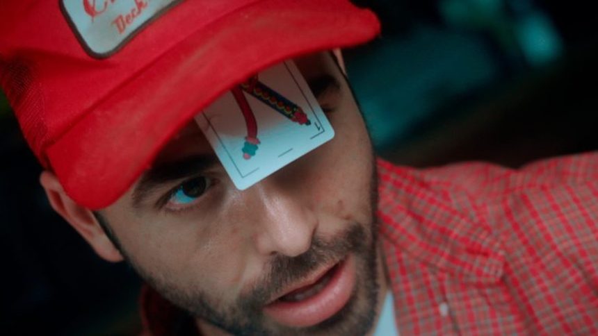 Homem com boné vermelho e carta de baralho no rosto, em evento de cinema.
