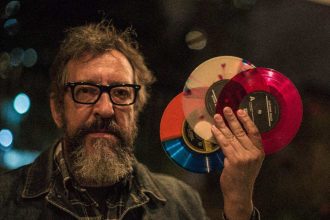 Homem com óculos e barba segura discos de vinil coloridos em evento musical.
