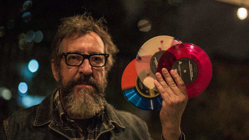 Homem com óculos e barba segura discos de vinil coloridos em evento musical.