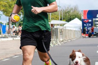 Homem correndo com cachorro em uma corrida, segurando bola e fazendo sinal de positivo.