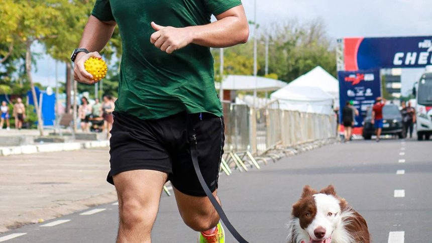 Homem correndo com cachorro em uma corrida, segurando bola e fazendo sinal de positivo.