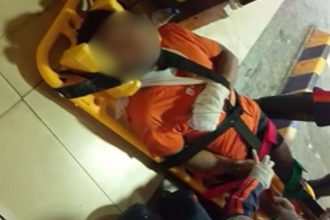 Homem ferido em maca com curativo no braço, possivelmente vítima de agressão.