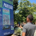 Homem usando smartphone para escanear código QR em painel de informações 'Paredão' em Manaus, Amazonas.