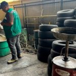 Homens em borracharia com pneus empilhados e tambores. Um deles tira fotos.