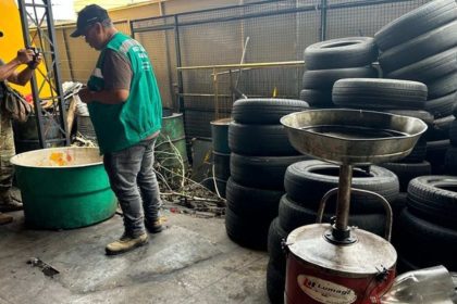 Homens em borracharia com pneus empilhados e tambores. Um deles tira fotos.