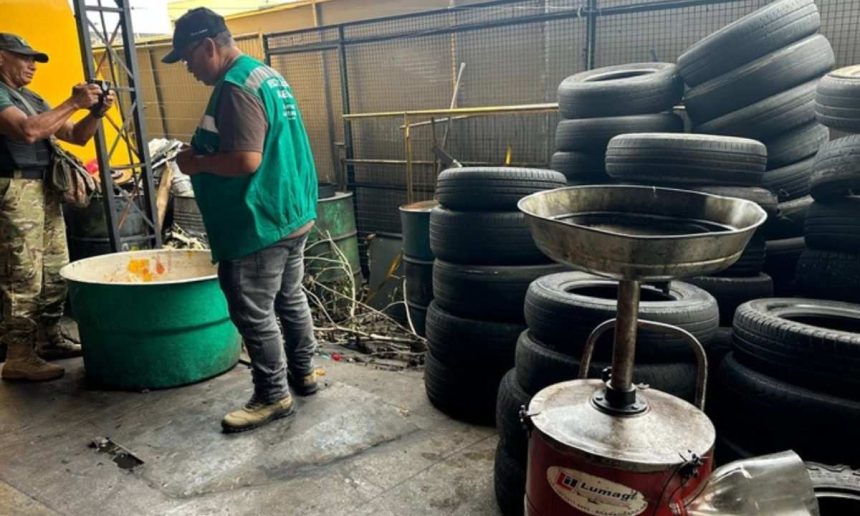 Homens em borracharia com pneus empilhados e tambores. Um deles tira fotos.