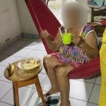 Idosa em rede comendo, simbolizando a situação de fome e abandono. Há comida em uma mesa próxima.