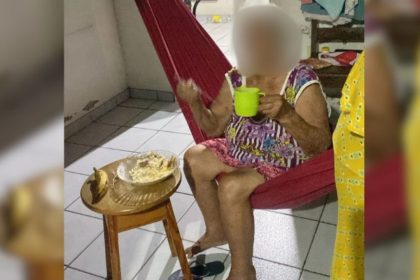 Idosa em rede comendo, simbolizando a situação de fome e abandono. Há comida em uma mesa próxima.
