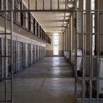 Interior de cela de prisão com grades de metal, corredor e luz no fundo