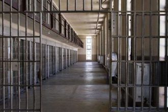 Interior de cela de prisão com grades de metal, corredor e luz no fundo