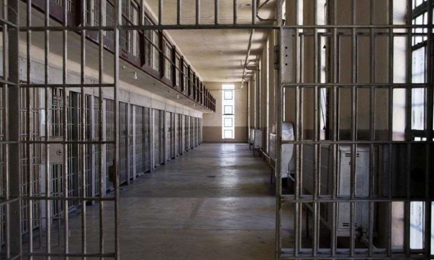 Interior de cela de prisão com grades de metal, corredor e luz no fundo