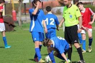 Jogadores de futebol em campo com árbitro. Um deles está no chão, próximo à bola.